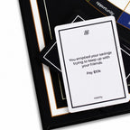 A white card with black text reading 'You emptied your savings trying to keep up with your friends. Pay $10k' and the word 'liability' at the bottom, placed on top of a black card deck with gold and white borders.