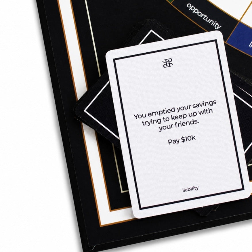 A white card with black text reading 'You emptied your savings trying to keep up with your friends. Pay $10k' and the word 'liability' at the bottom, placed on top of a black card deck with gold and white borders.